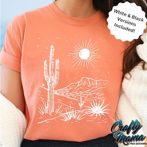 May include: Coral-colored t-shirt featuring a white desert landscape design with a cactus, sun, and mountains. The text "White & Black Versions Included!" is also visible. The shirt is a comfortable, casual style.