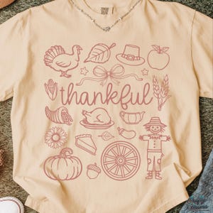 May include: Cream-colored t-shirt with a Thanksgiving-themed design in a muted rose color. The design includes a turkey, leaf, hat, apple, bow, cornucopia, roasted chicken, pie, pumpkin, and the word "thankful".