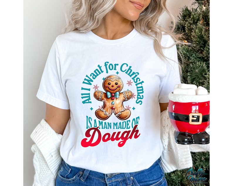May include: White t-shirt with a Christmas-themed graphic. The design features a gingerbread man and the text "All I want for Christmas is a man made of Dough". A Santa-themed mug is visible on the right.