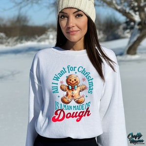 May include: White sweatshirt with a gingerbread man graphic and the text "All I want for Christmas is a man made of Dough". The gingerbread man has a blue bow tie and is surrounded by teal text and red text.