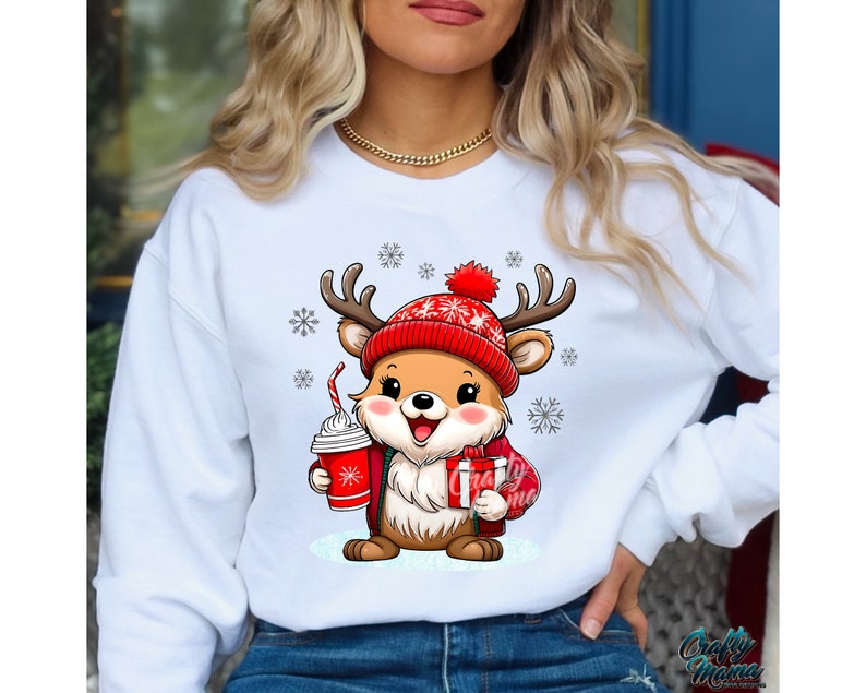 May include: White sweatshirt with a cute cartoon reindeer wearing a red hat and holding a cup of hot chocolate and a gift. The reindeer is surrounded by snowflakes.