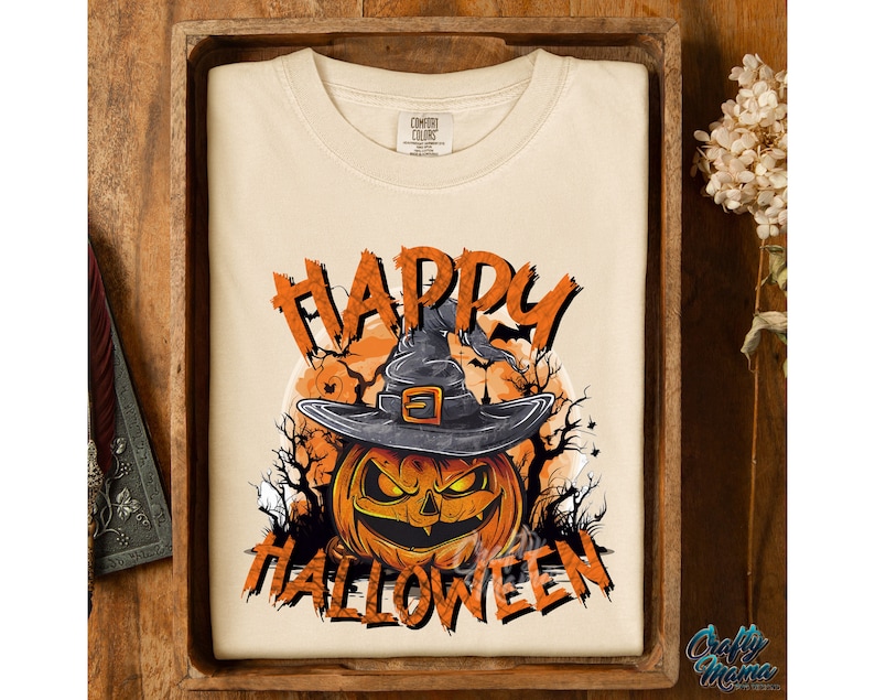 Peut inclure: Un t-shirt cr&egrave;me avec un motif de citrouille portant un chapeau de sorci&egrave;re et le texte "Happy Halloween".
