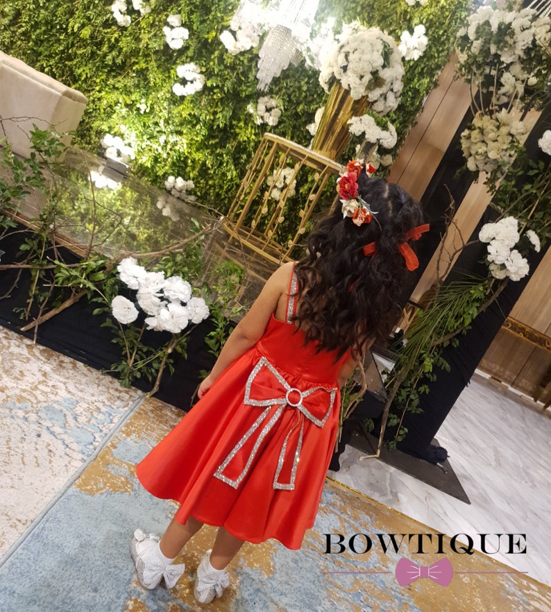 May include: A young girl wearing a red dress with a large, sparkly bow on the back. The dress has a satin finish and is decorated with silver sequins. The girl is wearing a flower crown and has long, curly hair.