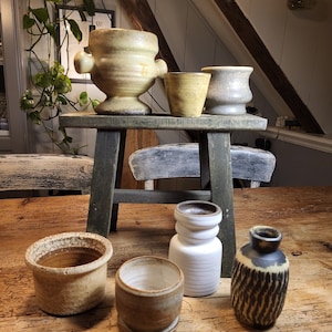 Könnte beinhalten: Eine Sammlung von sechs Keramikbehältern in verschiedenen Formen und Größen. Die Behälter sind in einer Mischung aus braunen, weißen und schwarzen Glasuren gehalten. Die Behälter stehen auf einem Holzhocker.