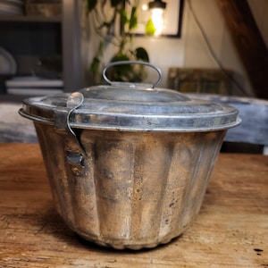 May include: A vintage silver-colored metal pudding mold with a fluted design and a hinged lid with a handle.