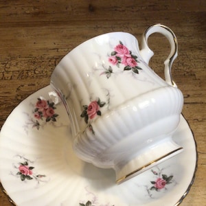 Pode incluir: Uma chávena de chá e um pires brancos com um delicado padrão floral com rosas cor-de-rosa e folhas verdes. A chávena tem uma borda dourada e uma pega. O pires tem uma borda ondulada e uma borda dourada.