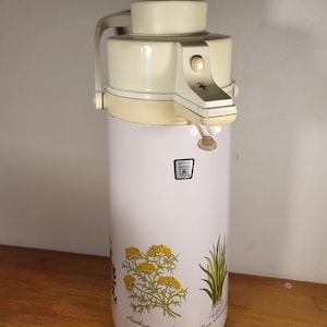 Könnte beinhalten: Vintage cremefarbene Thermoskanne mit weißem Korpus und botanischen Illustrationen. Der weiße Korpus zeigt gelbe Blumen und grüne Pflanzen. Die Thermoskanne hat einen cremefarbenen Griff und einen Druckknopfmechanismus. Höhe ca. 30 cm.