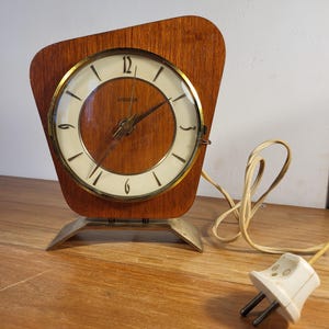 May include: A vintage wooden clock with a light-colored face and gold-colored trim. The clock has a unique, asymmetrical wooden frame and sits on a silver-colored metal base. A power cord with a white plug is visible.