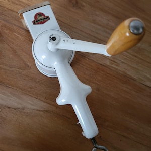 Puede incluir: Molino de alimentos blanco de manivela manual con mango de madera y manivela de metal. El molino tiene una etiqueta roja y blanca que dice "Röslehaus".