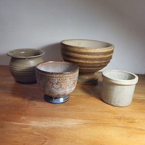 Op de afbeelding: Vier keramische potten in verschillende maten en kleuren. De potten zijn gemaakt van bruine, witte en grijze klei. Ze hebben een rustieke, handgemaakte uitstraling.