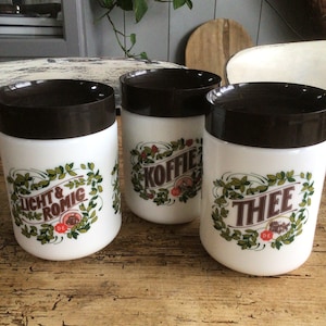 Op de afbeelding: Drie witte keramische potten met bruine deksels en groene en rode bloemenmotieven. De potten zijn gelabeld met "Licht & Romig", "Koffie" en "Thee".