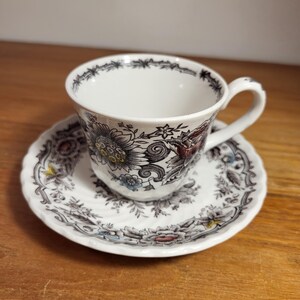 Op de afbeelding: Een wit theekopje en schotelset met een bloemenpatroon in tinten bruin, zwart, rood, geel en blauw.