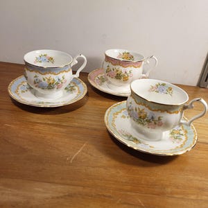 May include: Three white teacups and saucers with floral patterns. The cups and saucers have a pale blue border with gold edging. The cups have a curved handle and are on a wooden surface. The teacups and saucers are a set.