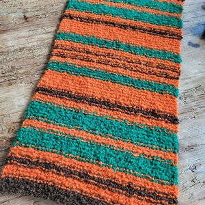 May include: A rectangular, hand-knitted rug with horizontal stripes of orange, teal, and brown. The rug has a textured, looped appearance and is likely made of a soft yarn. The rug is placed on a wooden surface.