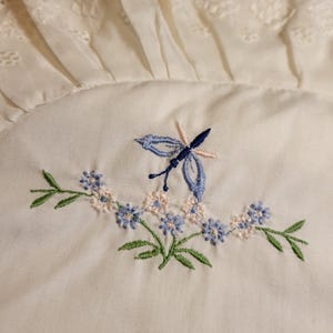 Peut inclure: Tissu blanc avec une broderie délicate de fleurs bleues et roses et un papillon bleu.