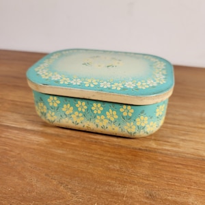 Pode incluir: Uma pequena caixa retangular de lata azul turquesa com uma tampa branca. A tampa tem um padrão floral branco e amarelo ao redor da borda e um desenho floral branco e amarelo no centro. A caixa está sobre uma superfície de madeira.
