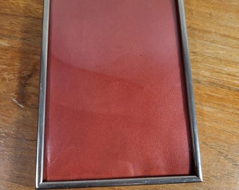 May include: A rectangular picture frame with a silver metal border. The frame contains a textured, reddish-brown surface. The frame is sitting on a wooden surface.