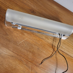 Op de afbeelding: Een vintage, rechthoekige tl-armatuur met een heldere, geribbelde plastic afdekking en chromen afwerking. Zwarte draden steken uit de armatuur. Het licht ligt op een houten oppervlak.