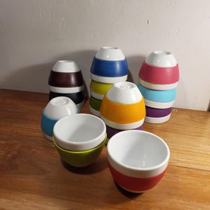 May include: A selection of colourful, stackable ceramic bowls. The bowls have a white interior and a variety of exterior colours, including blue, green, orange, pink, and brown. The bowls are arranged on a wooden surface.
