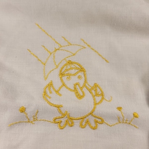Peut inclure: Tissu blanc avec une broderie jaune représentant un poussin sous un parapluie. Le poussin, le parapluie et la pluie sont brodés en fil jaune. L'ensemble donne un aspect artisanal.
