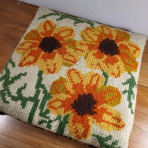 May include: Square throw pillow with a needlepoint design featuring three large sunflowers in shades of yellow, orange, and brown against a cream background. Green leaves and stems surround the flowers. The pillow is on a wooden surface.