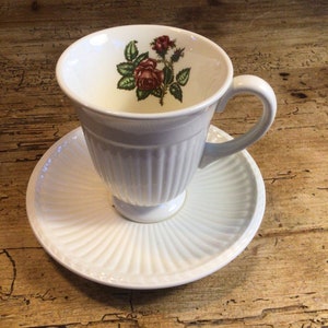 May include: White teacup and saucer with a floral design featuring a red rose and green leaves. The cup has a fluted design and a pedestal base.