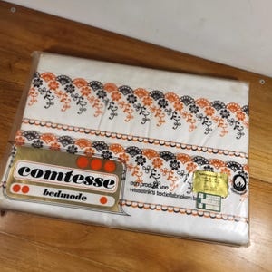 Pode incluir: Um lençol branco com um padrão floral preto e laranja. O lençol está embalado em plástico e tem um rótulo que diz "Comtesse Bedmode" e "een product van wisselink's textiel fabrieken b.v."