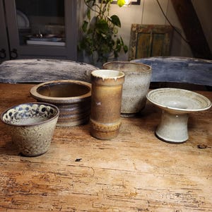 Könnte beinhalten: Fünf Keramikstücke in verschiedenen Formen und Größen. Die Stücke sind in Braun-, Beige- und Weißtönen mit gesprenkelten und strukturierten Oberflächen. Die Stücke sind auf einem Holztisch angeordnet.