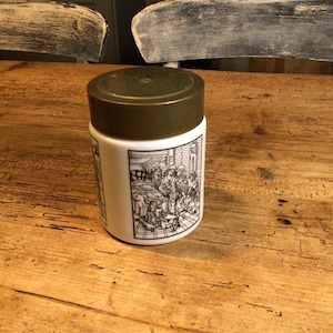 Op de afbeelding: Een witte keramische pot met een bruin deksel en een zwart-witte illustratie van mensen aan de zijkant. De pot staat op een houten tafel.