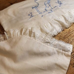 Peut inclure: Deux taies d'oreiller blanches avec des flamants roses brodés en bleu et une bordure en dentelle blanche. Les taies d'oreiller sont sur une surface en bois.