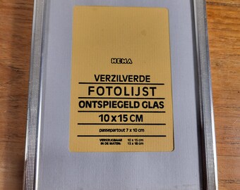 May include: A silver rectangular photo frame with a decorative edge. The frame contains a yellow label with black text, including "VERZILVERDE FOTOLIJST" and "10 x 15 CM". The frame is designed to hold a 10 x 15 cm photo.