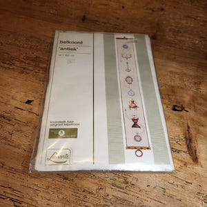 May include: A white and green cross stitch kit for a wall hanging. The kit includes a printed pattern, embroidery floss, and a needle. The pattern features a series of antique-style objects, including a clock, a teapot, and a candlestick. The kit is labelled "belkoord 'antiek'" and measures 14 x 100 cm.