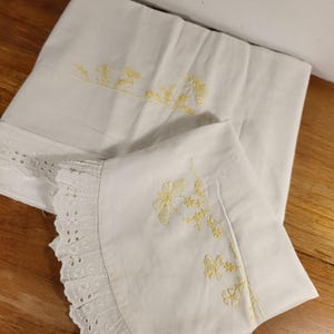 Peut inclure: Taies d'oreiller blanches avec broderie florale jaune. L'une des taies d'oreiller a une bordure décorative en dentelle ajourée. La broderie représente des papillons et des fleurs. Les taies d'oreiller sont pliées sur une surface en bois.
