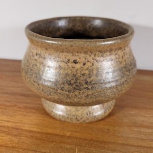 Puede incluir: Un cuenco con pedestal de cerámica moteada con un borde ancho y redondeado. El cuenco es de color marrón claro con motas marrones oscuras y un interior marrón oscuro. El cuenco se asienta sobre una base de pedestal corta y redondeada.