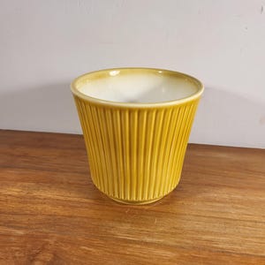 May include: A yellow ceramic plant pot with vertical ribbed detailing. The interior is a lighter cream colour. The pot sits on a wooden surface against a white wall. Suitable for indoor plants.