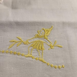 Peut inclure: Gros plan sur un tissu blanc avec une broderie jaune. La broderie représente une figure stylisée tenant des brins, avec une ligne pointillée. Le motif est probablement destiné à un textile décoratif.