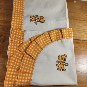 Peut inclure: Ensemble de literie crème avec bordure en vichy orange et motifs d'ours en peluche brodés. L'ensemble comprend un drap plat et un drap-housse, avec un bord à volants sur le drap-housse. L'ours en peluche est dans les tons orange et marron.