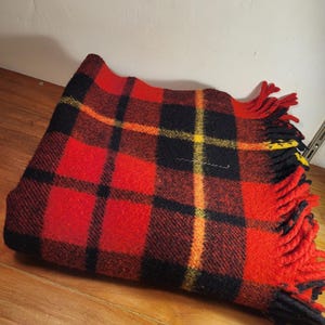 May include: A folded wool blanket featuring a vibrant plaid pattern in red, black, and yellow. The textile has a thick, textured weave and is finished with long, twisted fringe tassels along one edge, alternating in red, black, and yellow colours.
