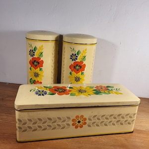 May include: Set of three vintage metal storage containers with a floral design. The containers are cream colored with yellow, orange, red, and blue flowers. The containers have a gold trim.