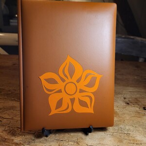 May include: A brown leather-bound album with a bright orange flower design on the front cover. The album is standing on a wooden table.
