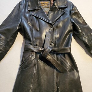 Peut inclure: Un manteau en cuir noir avec un design croisé, une ceinture à la taille et deux poches avant. Le manteau a un col classique et une étiquette qui dit "PALL MALL GEAR".