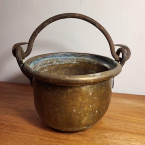 Puede incluir: Una pequeña olla de latón antigua con un asa de hierro oxidado. La olla tiene un fondo redondeado y un borde ligeramente acampanado. Es probable que se use para cocinar o servir.