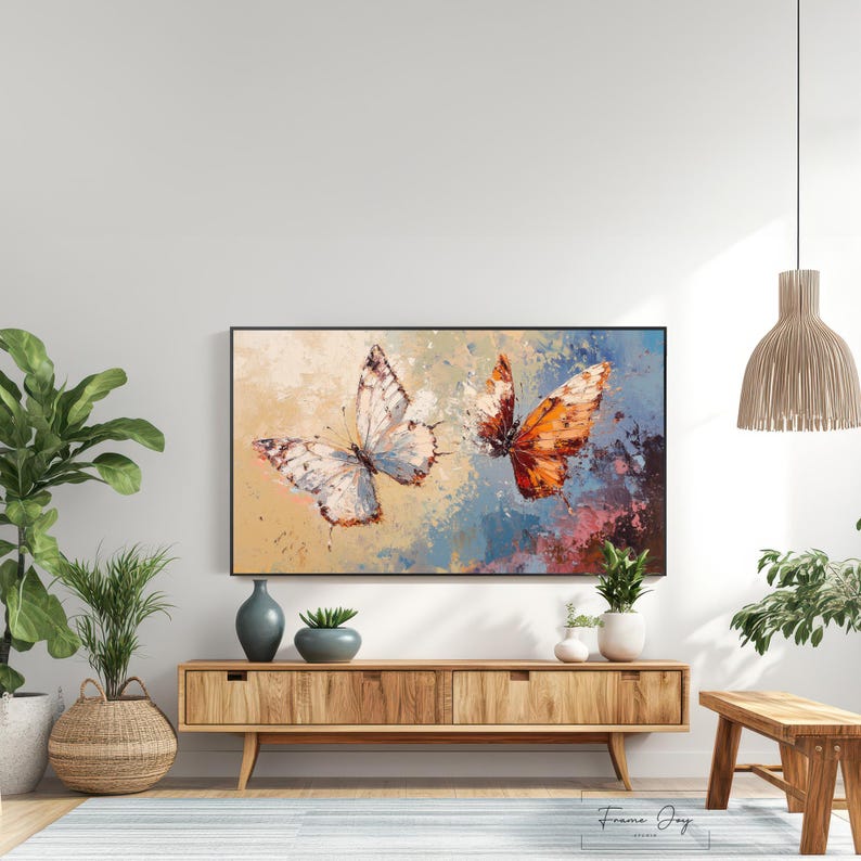 Op de afbeelding: Een ingelijst canvas kunstwerk met twee vlinders. E&eacute;n vlinder heeft witte en bruine vleugels, de andere heeft oranje en bruine vleugels. De achtergrond heeft een mix van blauwe, beige en roze kleuren. Het kunstwerk wordt boven een houten kast getoond.