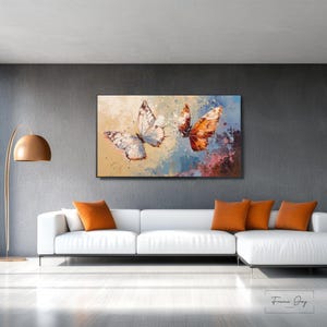 Op de afbeelding: Een ingelijst canvas kunstwerk met twee vlinders met gedetailleerde vleugelpatronen. E&eacute;n vlinder is wit met zwarte markeringen, de andere is oranje. De achtergrond heeft een mix van blauwe, gele en rode penseelstreken.
