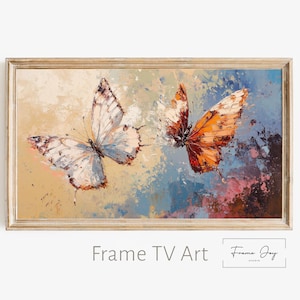 Op de afbeelding: Een ingelijst schilderij met twee vlinders. De ene vlinder heeft witte en blauwe vleugels, de andere heeft oranje vleugels. De achtergrond heeft een textuur, abstract ontwerp met beige, blauwe en roze tinten. De lijst is lichtbruin.