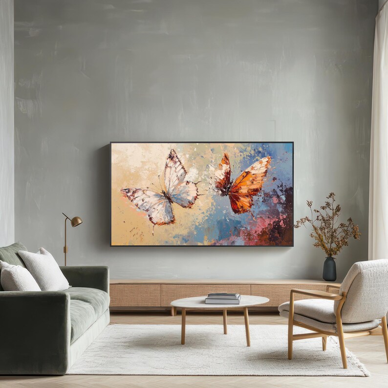 Op de afbeelding: Een groot, ingelijst canvas kunstwerk met twee vlinders. E&eacute;n is wit en grijs, de andere oranje en bruin. De achtergrond heeft een mix van blauwe, beige en roze tinten, wat een abstract effect cre&euml;ert. Het kunstwerk wordt tentoongesteld boven een lage houten kast.