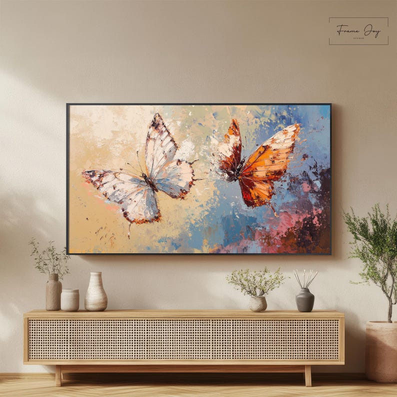 Op de afbeelding: Een ingelijst canvas kunstwerk met twee vlinders. E&eacute;n vlinder heeft witte en blauwe vleugels, de andere heeft oranje en bruine vleugels. De achtergrond heeft een mix van kleuren, waaronder blauw, geel en roze. Het kunstwerk wordt boven een houten kast getoond.