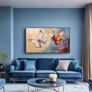 Op de afbeelding: Een ingelijst canvas kunstwerk met twee vlinders. E&eacute;n is wit, de andere oranje, tegen een achtergrond van beige, blauwe en bruine penseelstreken. Het kunstwerk hangt boven een blauwe bank.