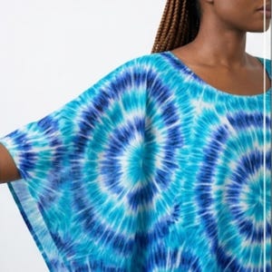 May include: A loose-fitting top featuring a vibrant tie-dye pattern in shades of bright turquoise, deep royal blue, and white. The garment has a wide, flowing sleeve design and a rounded neckline, showcasing a circular, radiating sunburst motif.