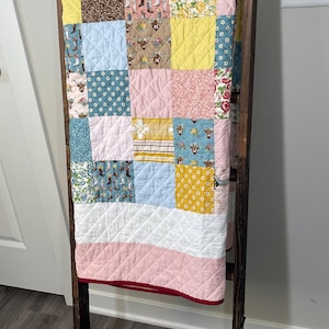 May include: A patchwork quilt featuring a variety of fabric squares in shades of yellow, pink, light blue, and white. The quilt displays patterns including floral prints, cowboy boots, and highland cattle, all finished with a diamond-stitched texture.